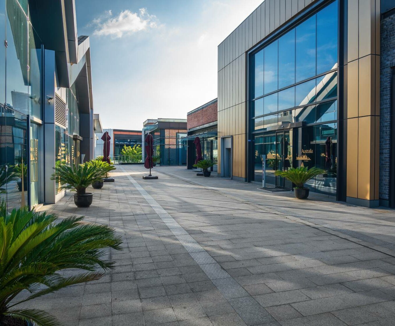 Modernt shoppingtorg med skyltfönster i glas och krukväxter längs en bred, tom gångväg under en klar himmel.